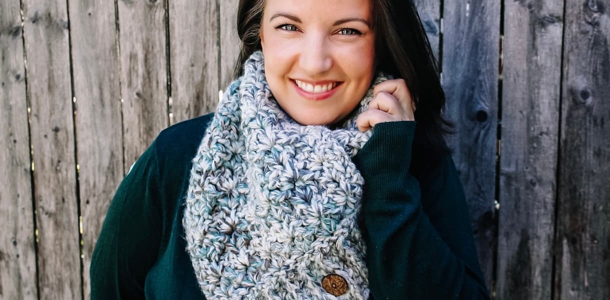 Smiling dark haired woman wearing dark green sweater, with green and cream seaglass crochet cowl with wooden buttons
