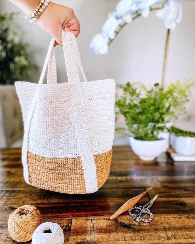 Mirabeau crochet tote bag in cream and mustard, held over a coffee table with yarn and hook nearby