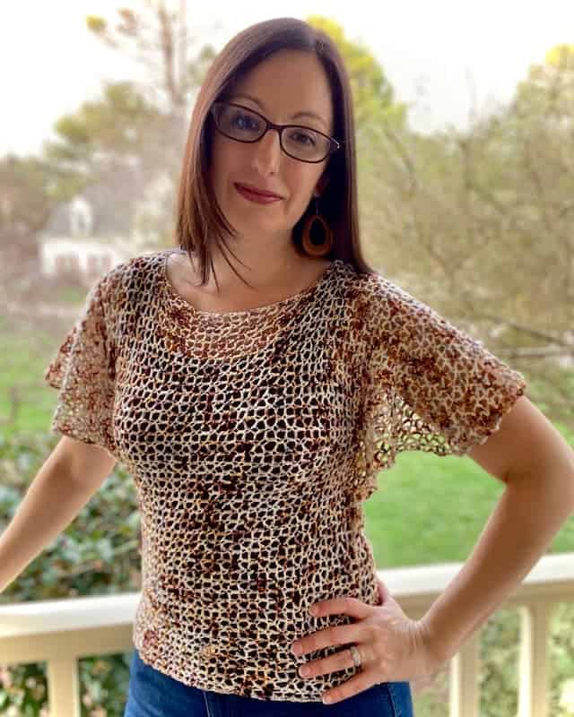 Dark haired woman wearing cream and brown lacy crochet top with flowing sleeves, leaning against a porch railing