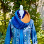 Blue and gold crochet infinity scarf with stripes fading from one colour to another, double around the neck of a mannequin wearing a blue denim jacket
