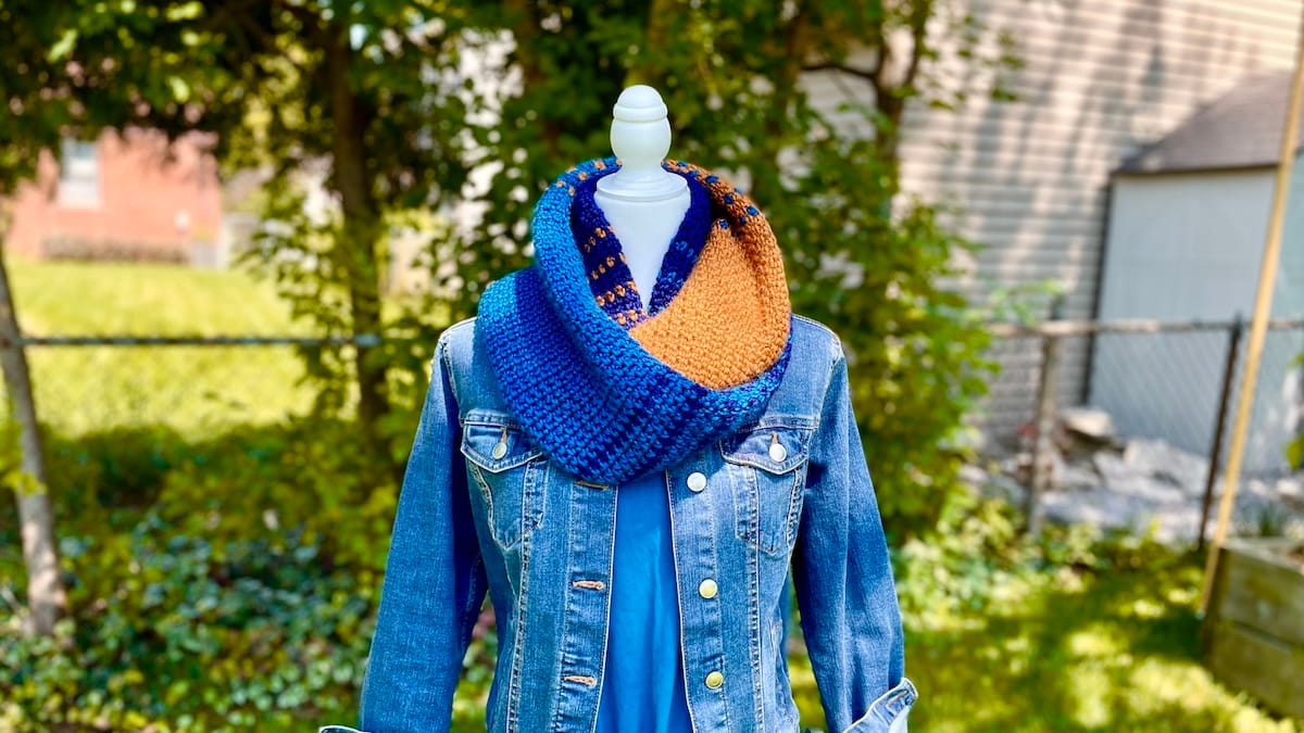 Blue and gold crochet infinity scarf with stripes fading from one colour to another, double around the neck of a mannequin wearing a blue denim jacket