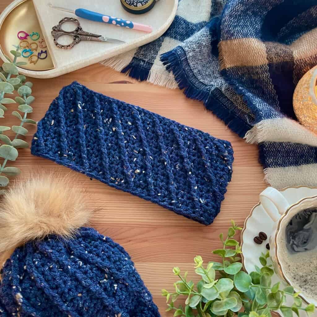Overhead view of the Cable Swirl crochet headband in dark blue on a table, showing the matching beanie pattern in bottom corner.